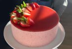 A beautiful, three-layered strawberry mousse cake on a white plate, with fresh strawberries and a dusting of powdered sugar on top