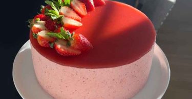 A beautiful, three-layered strawberry mousse cake on a white plate, with fresh strawberries and a dusting of powdered sugar on top