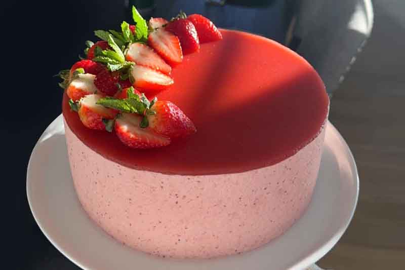A beautiful, three-layered strawberry mousse cake on a white plate, with fresh strawberries and a dusting of powdered sugar on top
